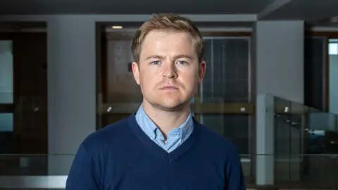 Queen's University Belfast A man with short blonde hair and blue eyes is looking directly into the camera, unsmiling. He is wearing a light blue shirt under a dark blue v neck jumper. The background is out of focus, but he is standing in the hallway of an office building. 