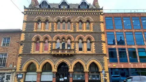 LDRS A historic brick building with decorative arches and patterned stonework stands along a town street, with bicycles parked outside and a modern building beside it.