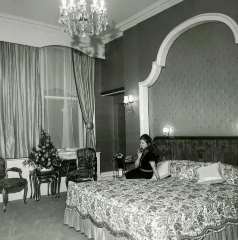 Grand Hotel A black and white image shows a woman sitting on a huge bed covered in a floral bedspread. She is talking on a telephone that is on a table next to the bed. 