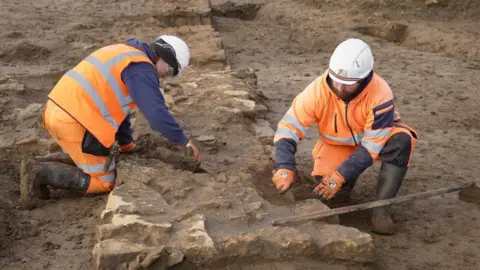 Two men, both dressed in high visibility clothing, are in a field leaning over the remains of a Roman wall. You can see the brickwork which looks like the base of a wall.
