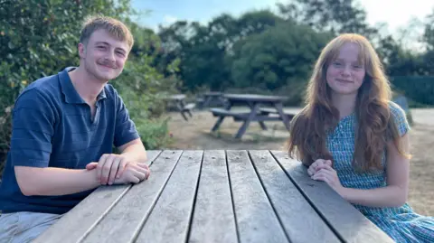 Bill Nicholson is wearing a blue top and sitting at a picnic table opposite Amber Thomson who is wearing a blue dress.