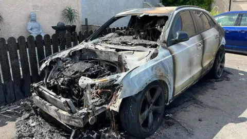BBC A burnt out silver car. The bonet has been removed showing fire damage inside. The side of the doors are burnt. It is in front of a brown fence.