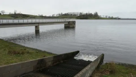 A reservoir on a grey day with green grass around it, a concrete pier and a boat dock are on the edge. 