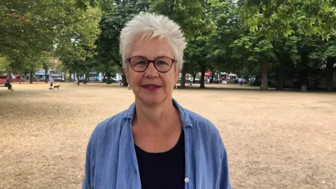 Liz Bodycote with short white hair, purple-framed glasses and a blue linen shirt stands on dried-out grass in a park, with trees and shops behind her.