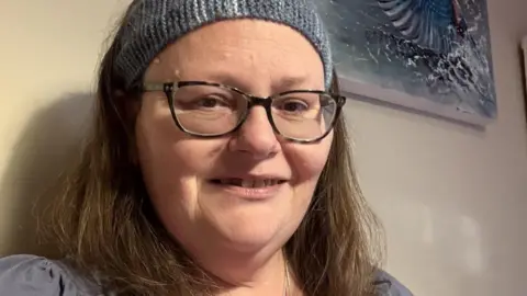 Supplied Selfie of Sarah Bodger smiling, wearing a blue headband, glasses, a necklace and a blue top.