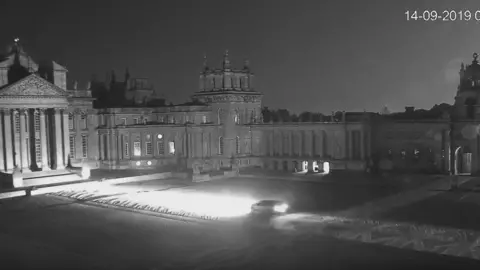 Thames Valley Police A night CCTV image of Blenheim Palace courtyard showing a car driving through it with headlights on. The ground leading up to the palace is overed in large Union flags