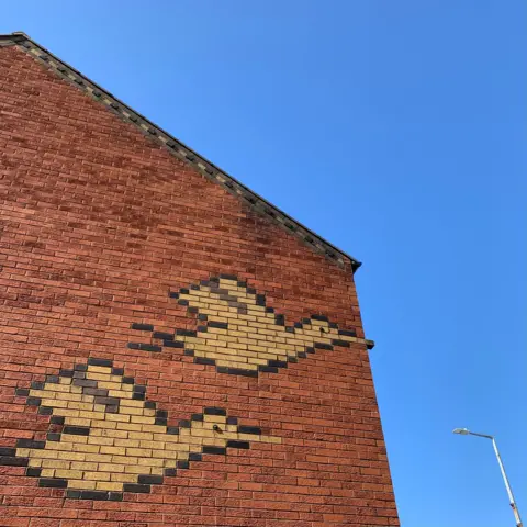Tom Hicks The red brick side of a building with the images of two flying ducks in a lighter coloured brick. They are outlined in a dark brick. The duck's bills are represented by two bricks, with the top duck's beak jutting out from the building against the blue sky 