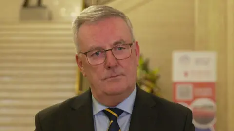 Mike is standing inside the Great Hall in Stormont beyond the steps. He has grey short hair and square glasses. He is wearing a blue shirt, navy and yellow tie and a black blazer jacket. 