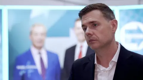 Matt Goodwin, in a open neck shirt and dark suit jacket, wears a serious expression. A large Nigel Farage poster is in the background