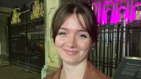 A girl with brown hair stands in front of city hall, she has on a brown jacket and gold necklace