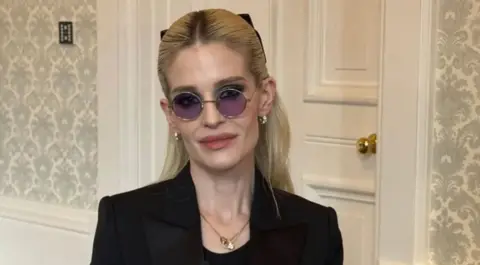 Kelly pictured slightly smiling while wearing purple lens circular glasses and a black blazer. She has blonde hair scraped back away from her face.