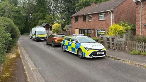Police cars parked on a residential street. They are outside a house with a boarded up window or door.
