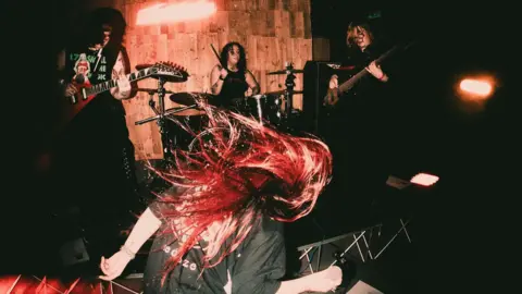 A woman with red hair is pictured rocking her head back and forth. Other band members holding guitars and playing drums can be seen in the background.