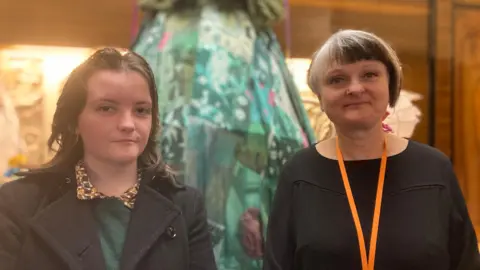 Michael and Stephanie Howell stand together in front of the puppet. Michael has long brown hair and is wearing a green jumper and black coat. Stephanie has short brunette hair with a fringe and is wearing a black top and orange lanyard.