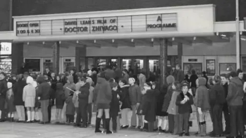 A black and white photograph showing a long line of people queuing to get into the old Odeon cinema when it was open.
