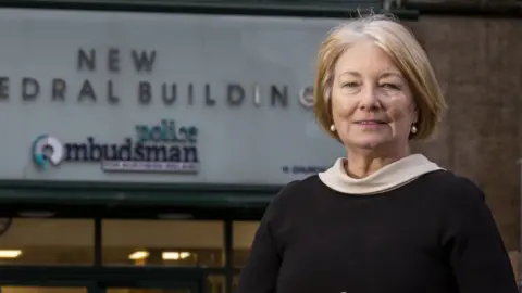 Marie Anderson outside her office in Belfast. She has one hand resting over the other and has a ring on both hands. She's wearing a silver watch and peral earrings and a black and white dress. The sign in the background says new cathedral building, police ombudsman.