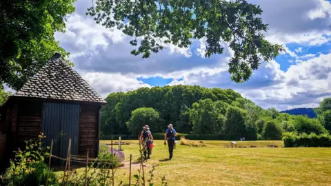 BBC Weather Watchers/ Margaret Grant Two people walking across some grass. There is a shed to the left and trees to the right.