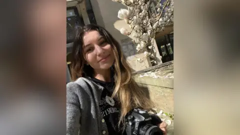 Iaroslava Photography Photographer Iaroslava smiling for a photo outside. She is holding a camera and has a black T-shirt with the logo of Oxford Shorts under a long-sleeve grey jacket. A magnolia tree is blooming next to her. It is sunny.