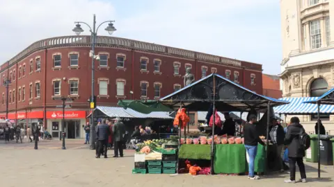 Market stalls selling fruit and veg in Walsall town centre, with people standing around, and buildings with shops and a bank in the background