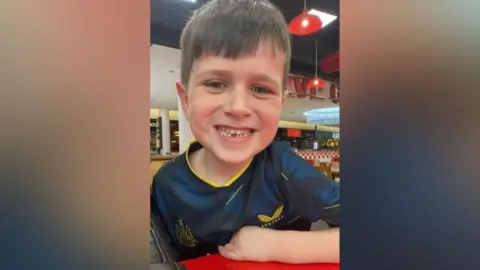 Archie York, a seven-year-old boy with brown hair, smiles at the camera. He is wearing a dark blue Newcastle football shirt.