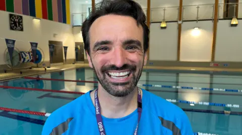 BBC/Lucinda Adam Russell has short black hair and a black bears. He stands smiling a swimming pool wearing a swimming teacher uniform and lanyard. 
