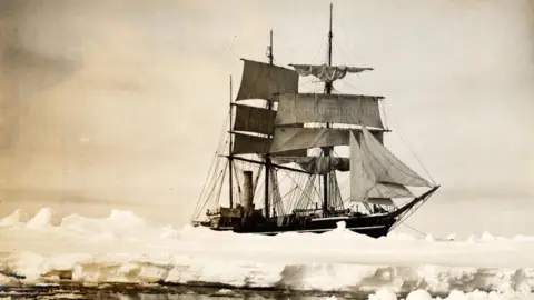 Getty Images/Royal Geographical Society Black and white image of the Terra Nova ship sailing in Antarctica. The ship is wooden, it's sails are open. It is moving across ice-covered ocean. 