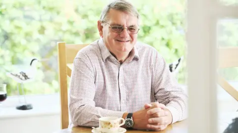 Photo of David oar smiling and sitting at a table. He is a middle-aged man with glasses and a lightly coloured checked shirt.
There is a cup and saucer on the table.