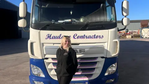 Ellie Shores Ellie Shores standing in front of a white and blue lorry with blue writing on it, reading "Haulage Contractors". It is parked in a yard. The sky is blue. She is wearing black trousers and a black hoodie with a round logo on. She has shoulder-length blonde hair and is wearing sunglasses on her head.