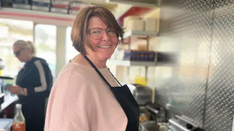 Rhona Hewit, a woman with brown hair and tortoiseshell glasses. She is working in a commercial-style kitchen, and wearing a light pink sweater and black apron. She is cooking on a flat-top griddle, with food items sizzling on the surface. In the background, another person is busy at a stainless steel counter. The kitchen is equipped with metal shelves stocked with containers and utensils.