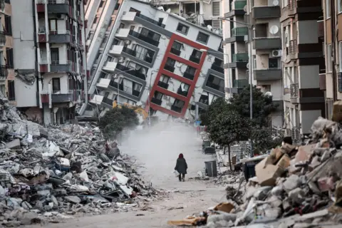 Sedat Suna / EPA A woman walks in front of a collapsed building while demolition teams work after a powerful earthquake in Hatay, Turkey