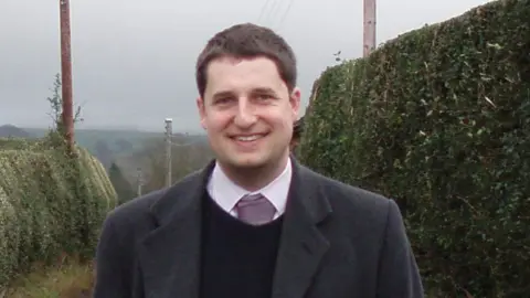 Supplied A tall man wearing a grey coat and a purple tie smiles at the camera. He is standing in a country lane, flanked by hedges.