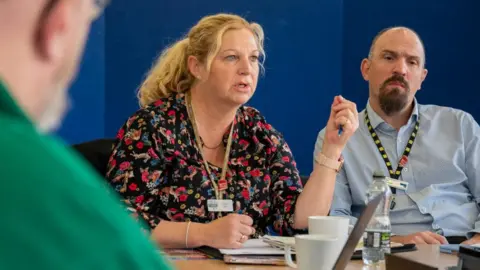 Northamptonshire OPFCC Alison Farr, wearing a floral top, with a lanyard round her neck, with one arm up, talking. She is sitting next to a man wearing a blue shirt, with a lanyard round his neck. She is sat in front of a blue background. A man in a green top is blurred and to the left. 