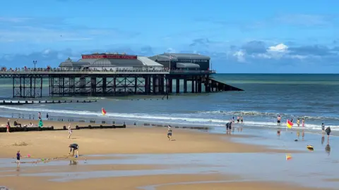 A wide view of people playing on a beach on a sunny day. There is a pier in the distance with a large building positioned at the end of the structure. 