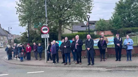 Kate Bradbrook/BBC A line of people of various ages standing on a pavement in front of a railing on which there is a light blue banner. There are trees behind the railing. Houses are visible to the right. There is a red sign banning lorries from proceeding.