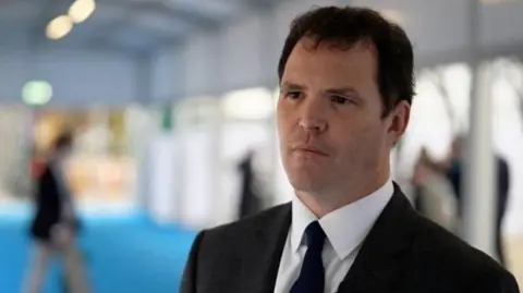 The head and shoulders of Lee Waters, who is stood at the right of the picture, can be seen in the picture. He is wearing a black suit and tie, and a white shirt. He is stood in a building which is out of focus behind him.