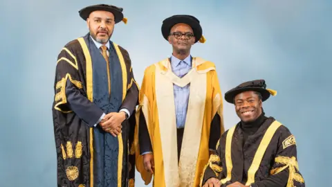 Birmingham City University Dr Vanley Burke, dressed in a gold graduation gown stood in the middle of two men. One is standing and one is sitting in a wheelchair.