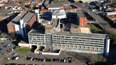 An aerial view of Anglia Square. Cars are parked outside a concrete and glass building. A road is situated to the left of the building and homes are in the background