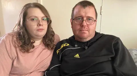 Paula Reece and Simon Hunt, both wearing glasses, sitting down facing the camera. Ms Reece wears a salmon coloured top and Mr Hunt a black Adidas hooded top.