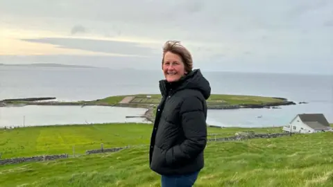 Ingrid Sutherland Ingrid Sutherland is wearing a black winter jacket and is looking at the camera. Behind her is the sea and a small green island. On her right is a small white house. 
