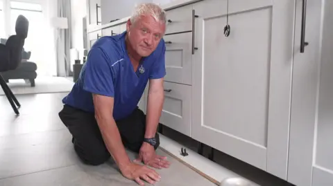 A man with short white hair wearing a blue T-shirt and black trousers kneels on the ground in a white kitchen. He is looking directly into the camera.