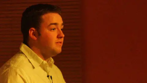 Stuart Steele/Civic Comedy A clean shaven man with dark hair, wearing a white shirt is looking to the right. He is against a red background