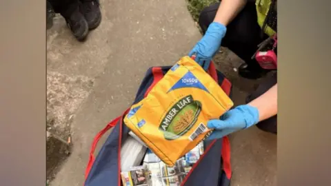A police officer wearing blue rubber gloves holds open a large blue sports bag that is packed with counterfeit tobacco. 