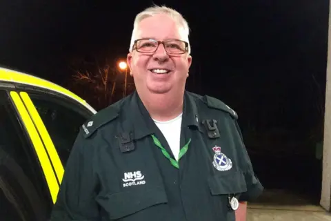 Clare Moore A smiling paramedic standing in front of an emergency service vehicle. He is wearing a green NHS Scotland uniform over a white t-shirt, has rectangular glasses and short white hair.