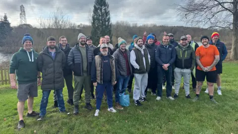 A group of men gather on a patch of grass next to a lake, which can be seen in the background.