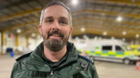 Man with a beard wears a green paramedic uniform and is photographed standing in a large warehouse with ambulances in the background.