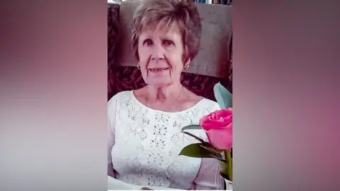 Maureen Sanderson, wears a white top and is smiling slightly. A pink flower is in the fore front of the right hand side of the picture.