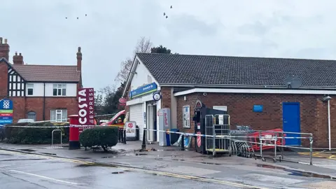 Sarah-May Buccieri / BBC The Co-op store in Billinghay. It is a small, single-storey brick building with a pitched roof. At the front is a forecourt with a tree and bushes and a red post box and a blue Co-op sign listing the services. Police tape surrounds the shop and the white window shutter is down.