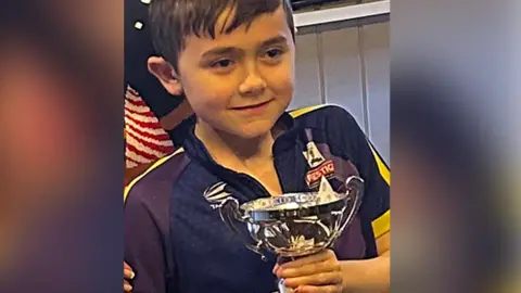 Family photograph 11-year-old Harry with short brown hair wearing a purple and yellow Luke Littler darts shirt clutching a silver trophy he won at a darts competition. He has a huge smile.