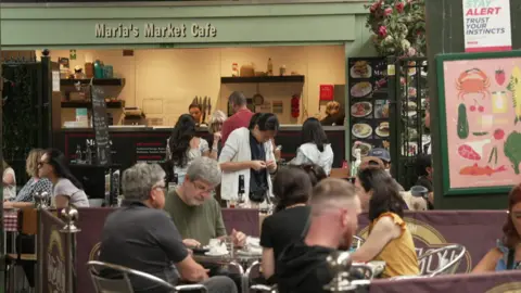 Thomas Magill/BBC A candid shot shows diners seated at tables outside a cafe. The cafe counter can be seen, under the cafe frontage which is pale green and has a sign reading "Maria's Market Cafe" in white lettering.