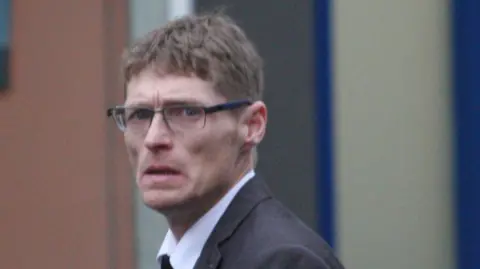 Central Scotland News A skinny man with brown hair and glasses. He looks worried and is wearing a suit. 